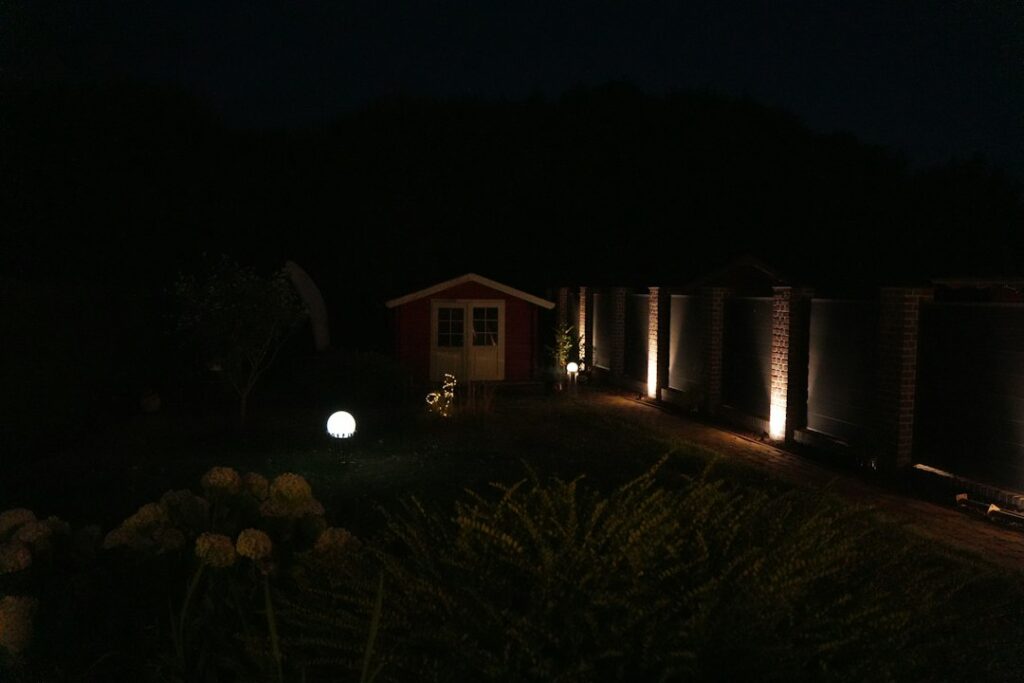 Warm outdoor lights illuminate a backyard path, patio seating, trees, and a pool edge after sunset.